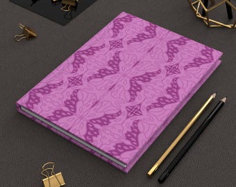 May include: A closed journal with a purple floral pattern cover. The journal is surrounded by gold binder clips, a black and gold pencil, and a gold pencil. The journal is on a dark gray surface.