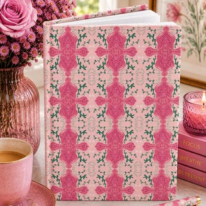 May include: A hardcover journal featuring a repeating pattern of pink abstract shapes and green vine-like motifs on a light pink background. The book has white pages and is accompanied by a matching pink pen with a gold-colored tip and clip.
