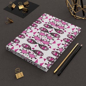 May include: A hardback notebook featuring a white cover with a repeating pattern of pink and black abstract shapes, including paisley-like motifs and small triangles. The notebook is accompanied by a gold-toned pen and a black pen.