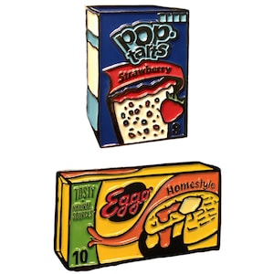 May include: Two enamel pins featuring popular breakfast food brands. The first pin is a blue and red Pop-Tarts box with the text "Pop-Tarts Strawberry" and a picture of a strawberry Pop-Tart. The second pin is a yellow and orange Eggo box with the text "Eggo Homestyle" and a picture of a waffle.