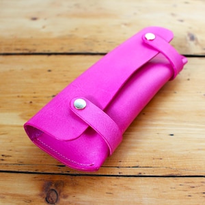 May include: A bright pink leather pencil case with a snap closure and a strap. The case is lying on a wooden surface.