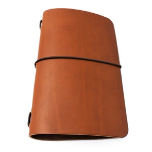 May include: A brown leather notebook with an elastic closure. The notebook is closed and lying on a white surface.