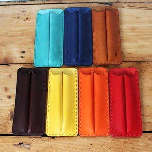 May include: Eight leather pen cases in different colours, including teal, navy blue, brown, black, yellow, orange, and red. Each case has two slots for pens and is stitched around the edges.