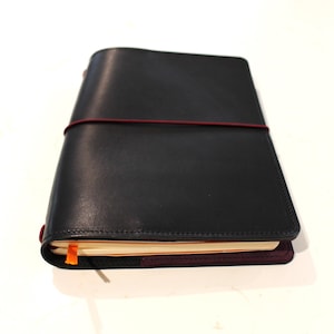 Puede incluir: Una funda de cuero negro para cuaderno con un cierre de banda elástica de color rojo oscuro que envuelve horizontalmente el exterior. La funda contiene un cuaderno con páginas de color crema y un pequeño marcapáginas de cinta naranja brillante.