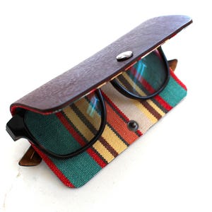 May include: A pair of black-framed spectacles resting inside a semi-open case. The case features a dark brown leather-effect top flap with a silver snap button and a lower section made of woven fabric with vertical stripes in teal, yellow, and red.
