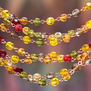 May include: A multi-strand necklace featuring faceted glass beads in shades of yellow, olive green, amber, and deep red, all linked together by a delicate copper-toned metal chain. The beads are arranged in several cascading, overlapping layers.