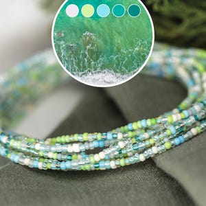 May include: Strands of colorful seed beads in shades of green, blue, white, and silver are draped over a green fabric. The beads are small and delicate, creating a textured effect. A color palette of ocean tones is in the upper left.