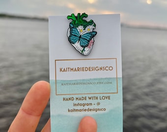 May include: A green enamel heart-shaped pin with a blue butterfly and white flowers. The pin is on a white card with the text "KAITMARIEDESIGNSCO" and "KAITMARIEDESIGNSCO.ETSY.COM". The card also says "HAND MADE WITH LOVE" and "instagram - @kaitmarie designsco".