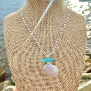 Pu&ograve; includere: Una collana a catena d'argento con un ciondolo a forma di conchiglia e un pezzo di vetro di mare blu turchese. La collana &egrave; esposta su un manichino di iuta.