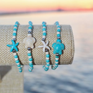 Puede incluir: Cuatro pulseras de cuentas con cuentas turquesas, blancas y plateadas. Cada pulsera presenta un dije diferente: una estrella de mar turquesa, una tortuga blanca, una estrella de mar blanca y una tortuga turquesa. Las pulseras se exhiben sobre una superficie de color neutro.