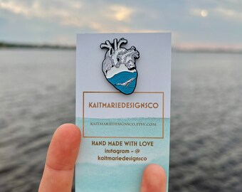 May include: White enamel pin of a heart with a blue ocean inside. The ocean has a white whale swimming in it. The pin is on a white card with the text "KAITMARIEDESIGNSCO" and "HAND MADE WITH LOVE Instagram - @kaitmarie designsco".