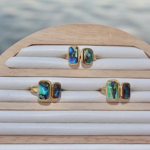 Puede incluir: Tres anillos de oro con incrustaciones de nácar de abulón iridiscente. Los anillos se muestran en un soporte de anillos de madera con forro de terciopelo blanco.