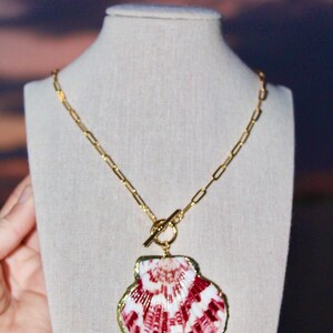 May include: A gold chain necklace with a large, red and white seashell pendant. The seashell is attached to the chain with a gold toggle clasp.