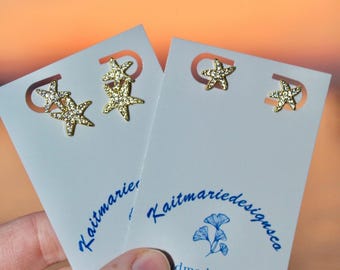 Puede incluir: Pendientes de estrella de mar dorados sobre tarjetas blancas. Un par presenta múltiples estrellas de mar, el otro una sola. Las tarjetas tienen el texto "Kaitmarie Designs Co" y "dmade Jewelry". El fondo es una puesta de sol borrosa.