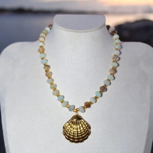 Peut inclure: Un collier de perles avec un pendentif coquillage doré. Le collier est composé d'une chaîne de petites perles colorées dans les tons de bleu, marron et blanc. Le pendentif est un coquillage texturé et détaillé, sur le thème côtier.