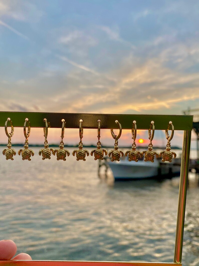 Op de afbeelding: Een display van gouden creolen met een klein schildpadbedeltje dat aan elke creool hangt. De oorbellen worden getoond tegen een achtergrond van een zonsondergang boven een waterpartij.