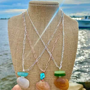 May include: Three necklaces with different colored sea glass and shell pendants. The necklaces are on a burlap mannequin and are layered on top of each other. The necklaces are silver, rose gold, and gold.