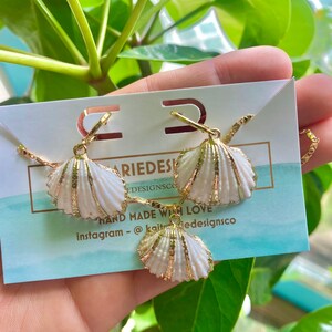 May include: A set of gold shell earrings and a necklace. The shells are white with gold accents. The jewelry is displayed on a white card with the text "Kait Designs Co. Hand Made With Love Instagram - @kaitdesignsco"