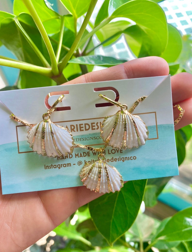 May include: A set of gold shell earrings and a necklace. The shells are white with gold accents. The jewelry is displayed on a white card with the text "Kait Designs Co. Hand Made With Love Instagram - @kaitdesignsco"