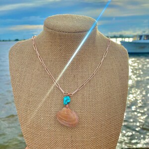 Pu&ograve; includere: Una collana con catena in rame con un ciondolo a forma di conchiglia e una pietra turchese. La collana &egrave; esposta su un manichino in iuta su uno sfondo di cielo blu e acqua.