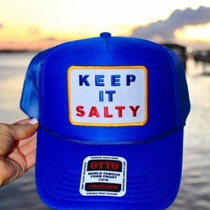 May include: A blue trucker hat with a white patch that reads "KEEP IT SALTY" in blue and red lettering. The hat has a mesh back and a black rope detail. The hat is made by Otto and has a durable snap closure.