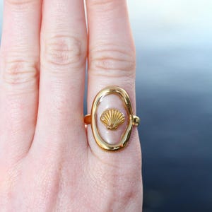 May include: A gold ring with a clear oval-shaped setting featuring a small gold seashell design.