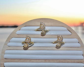May include: Gold-toned rings with a seashell design, adorned with small, sparkling stones. The rings are displayed on a wooden jewelry holder with white padded slots. The background features a sunset over the water.