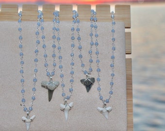 May include: Five necklaces with silver chains and blue glass beads, each featuring a different shark tooth pendant. The pendants are a mix of gray and white, and some have a wire wrap around the base.