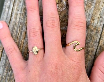 May include: Two gold rings on a hand. One ring features a seashell design, while the other ring is shaped like a wave.
