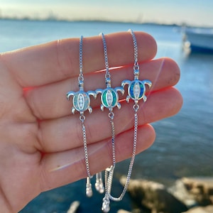 May include: Three silver turtle necklaces with adjustable chains. Each turtle pendant has a different coloured shell: white, green, and blue. The necklaces are shown against a blurred background of water and sky.
