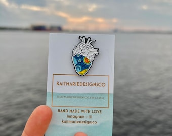 May include: A white enamel pin with a blue and yellow anatomical heart design. The pin is on a white card with the text "KAITMARIEDESIGNSCO" and "HAND MADE WITH LOVE" in black text. The text "Instagram - @kaitmarie designsco" is also on the card.