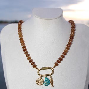 Peut inclure: Un collier de perles marron avec un fermoir doré et des breloques. Le fermoir retient une boussole, un coquillage turquoise et un petit poisson. Le collier est présenté sur un mannequin blanc. Les perles mesurent environ 0,5 cm de diamètre.