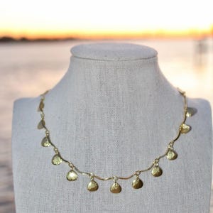 Può includere: Una collana color oro con piccoli ciondoli a forma di conchiglia. La collana è esposta su un manichino bianco. Lo sfondo mostra una vista sfocata di un tramonto sull'acqua.