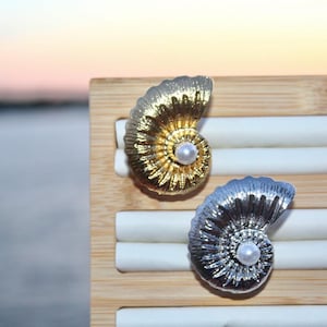 May include: Two gold and silver nautilus shell earrings with a pearl accent. The earrings are displayed on a wooden stand.