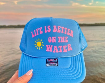 May include: A light blue trucker hat with a pink and yellow graphic that says "Life is better on the water". The hat has a black label that says "Otto World Famous Foam Front Trucker".