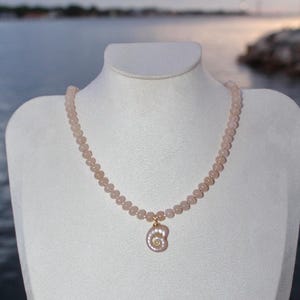 Peut inclure: Un collier délicat composé de petites perles roses pâles. Un pendentif en forme de coquillage, suspendu par une bélière dorée, orne le centre. Le collier est présenté sur un mannequin blanc, avec un arrière-plan extérieur flou.