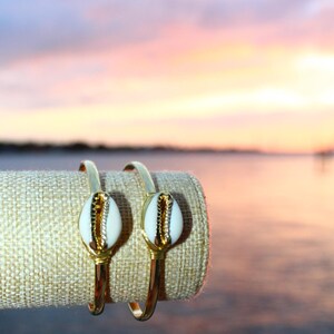 Może przedstawiać: Dwie bransoletki w kolorze złotym z białymi i złotymi akcentami muszli. Bransoletki są prezentowane na beżowym wałku z tkaniny na tle zachodu słońca nad wodą. Elegancka biżuteria o tematyce plażowej.