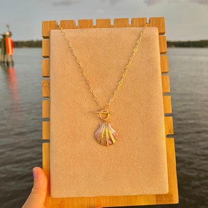 May include: A gold chain necklace with a pink and gold seashell pendant on a wooden display stand.