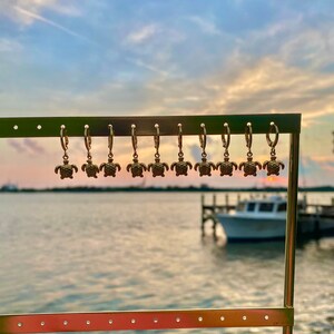Op de afbeelding: Een display van gouden creolen met een schildpadbedel die aan elke creool hangt. De oorbellen worden getoond op een metalen standaard tegen een wazige achtergrond van een waterpartij en een boot.