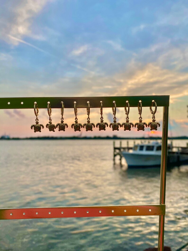 Op de afbeelding: Een display van gouden creolen met een schildpadbedel die aan elke creool hangt. De oorbellen worden getoond op een metalen standaard tegen een wazige achtergrond van een waterpartij en een boot.