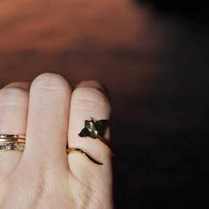 Puede incluir: Anillo dorado con diseño de hoja. El anillo se envuelve alrededor del dedo con una banda curva. Se ven anillos dorados adicionales. El fondo es una mezcla borrosa de naranja y negro.