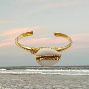 Può includere: Un bracciale a fascia in metallo dorato con una singola conchiglia di ciprea bianca al centro. La conchiglia è fissata alla fascia liscia e aperta con un sottile filo metallico dorato avvolto su entrambi i lati per un design coeso.