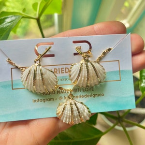 May include: A set of gold shell earrings and a matching necklace. The shells are white with gold accents. The jewelry is handmade with love and the Instagram handle @kmarie designsco is included on the card.