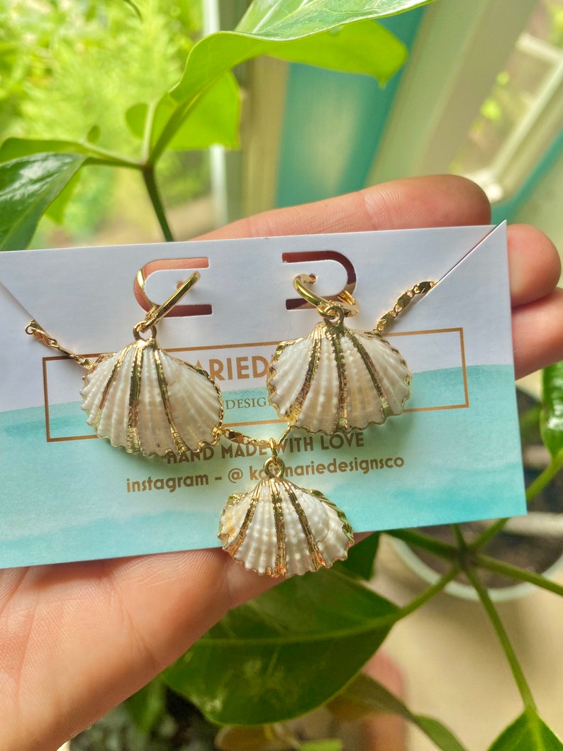 May include: A set of gold shell earrings and a matching necklace. The shells are white with gold accents. The jewelry is handmade with love and the Instagram handle @kmarie designsco is included on the card.