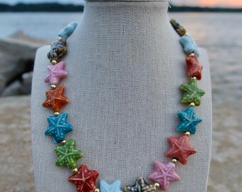 May include: A colorful starfish necklace displayed on a mannequin. The necklace features various colored starfish beads, including blue, green, red, and pink, strung together with gold-colored beads. The background shows a sunset over water.
