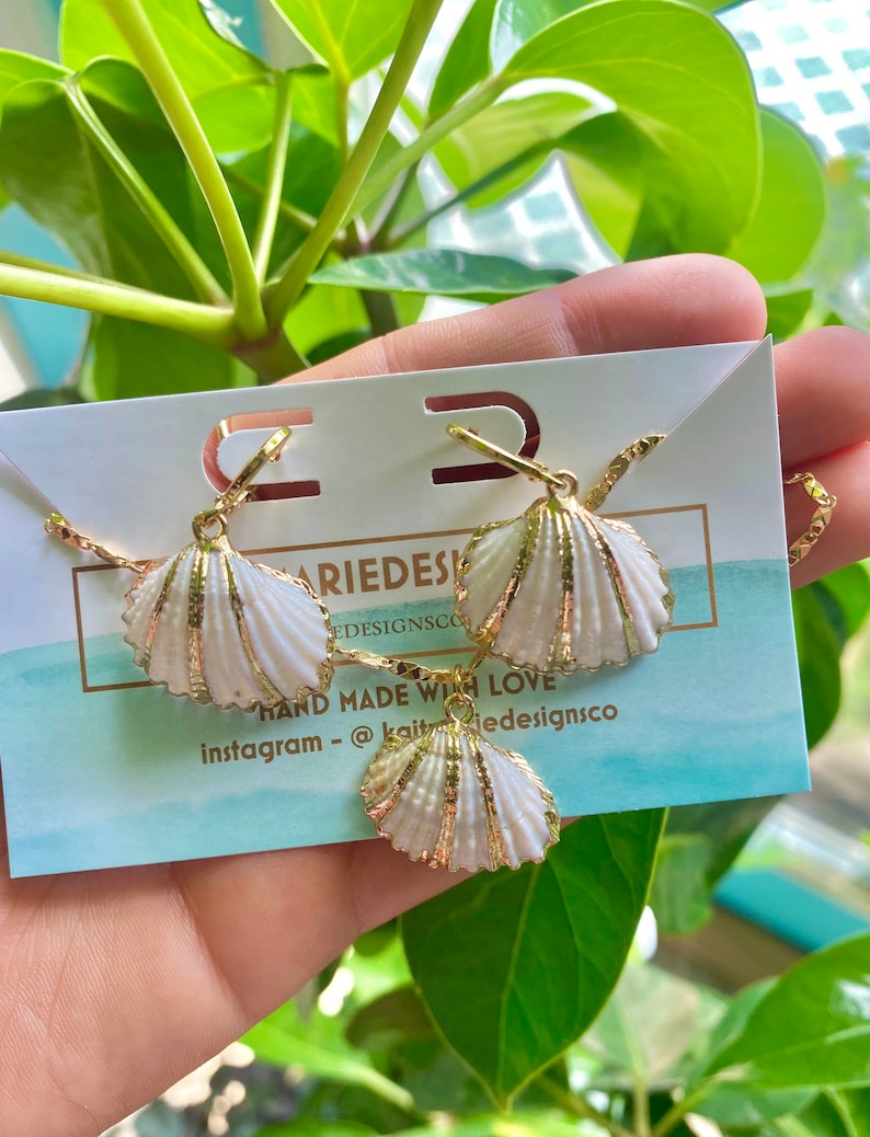 May include: A set of gold shell earrings and a necklace. The shells are white with gold accents. The jewelry is displayed on a white card with the text "Kait Designs Co. Hand Made With Love Instagram - @kaitdesignsco"