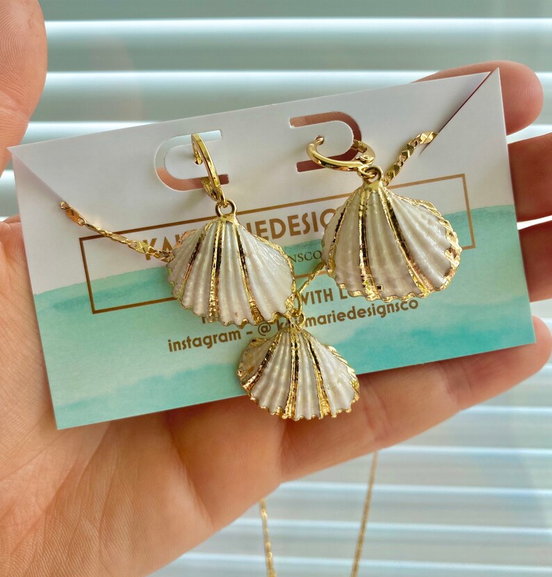 May include: A gold necklace and earrings set featuring white seashell charms. The necklace has a delicate chain and the earrings are hoop style. The set is displayed on a white card with the text "marie designsco" and "instagram - @marie designsco".