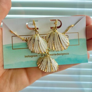 May include: A gold necklace and earrings set with white seashell charms. The necklace has a delicate chain and the earrings are hoop style. The charms are detailed with gold accents.  The text on the card reads "Handmade with love" and "instagram - @marie designsco".