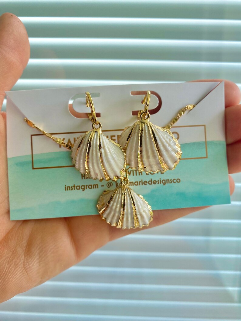 May include: A gold necklace and earrings set with white seashell charms. The necklace has a delicate chain and the earrings are hoop style. The charms are detailed with gold accents.  The text on the card reads "Handmade with love" and "instagram - @marie designsco".