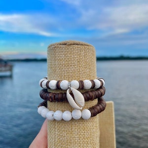 Könnte beinhalten: Drei Armbänder, die auf einem braunen Jute-Präsentationsständer gestapelt sind. Das obere Armband besteht aus weißen Lavasteinen mit einem kleinen weißen Muschel-Anhänger. Das mittlere Armband besteht aus braunen Holzperlen. Das untere Armband besteht aus weißen Lavasteinen.